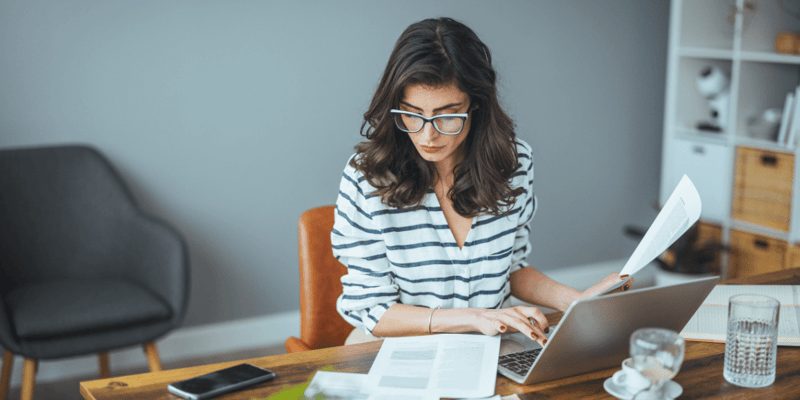 A woman in glasses working on a laptop at a wooden table with papers and a smartphone.
